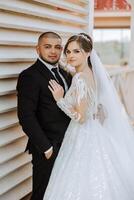 Stylish, young groom and beautiful bride in a long white dress and a long veil with a bouquet in their hands, hugging in the park in the autumn nature. Wedding portrait of newlyweds. photo