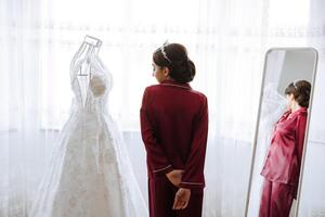 morning portrait of a luxurious and beautiful bride in a hotel in a luxurious room in red pajamas standing next to her wedding dress. photo