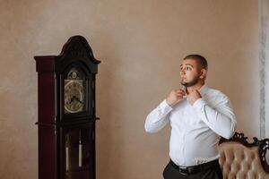 un hombre en un blanco camisa soportes por el ventana en el habitación y sujeta el botones en su collar y mangas reloj en mano. elegante negocio retrato de un hombre, de cerca foto. el novio es preparando. foto