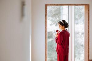 The morning of a luxurious and beautiful bride in a hotel in a luxurious room in red pajamas, standing by the window with a red rose in her hands. photo