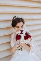 retrato. un morena novia en un vestir y velo, un elegante corona poses y olfatea su ramo. plata joyas. hermosa maquillaje y cabello. verano boda. celebracion foto