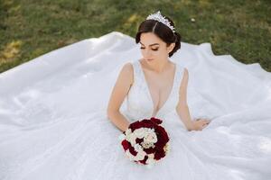 A brunette bride with a tiara in her hair, sitting on the ground in a dress unfolded in the shape of a circle, holding a bouquet. On a green background. Sunny day. Wedding ceremony photo