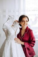 morning portrait of a luxurious and beautiful bride in a hotel in a luxurious room in red pajamas standing next to her wedding dress. photo