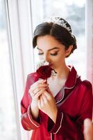 Vertical morning portrait of a luxurious and beautiful bride in a hotel in a luxurious room in red pajamas, standing by the window with a red rose in her hands. photo