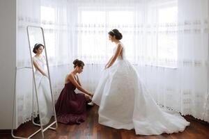 The bridesmaid is happy in the morning, helping to fasten the buttons on the wedding dress and prepare for the wedding ceremony. They take pictures, smile, help the bride with her shoes. photo