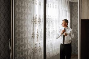 a young man in a white shirt stands by the window in the room and puts on a tie. The groom gets dressed in the morning and prepares for the wedding. photo