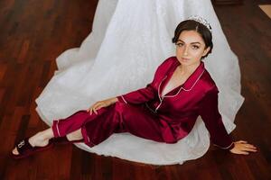 morning portrait of a luxurious and beautiful bride in a hotel in a luxurious room in red pajamas sitting and posing next to her wedding dress. photo