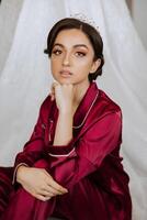 morning portrait of a luxurious and beautiful bride in a hotel in a luxurious room in red pajamas sitting and posing next to her wedding dress. photo