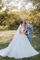 Stylish, young groom and beautiful bride in a long white dress and a long veil with a bouquet in their hands, hugging in the park in the autumn nature. Wedding portrait of newlyweds. photo