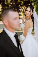 Portrait of a smiling bride with a bouquet of flowers hugging her husband while looking at the camera. Beautiful wedding celebration. Wedding hairstyle with a veil. Couple in love. photo