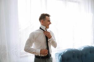 un joven hombre en un blanco camisa soportes por el ventana en el habitación y pone en un atar. el novio obtiene vestido en el Mañana y prepara para el boda. foto