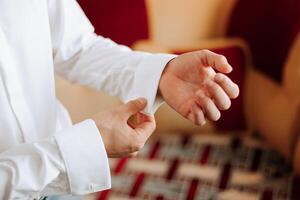 A man in a white shirt stands by a window in a room and fastens buttons on his sleeves, hands close-up. Watch on hand. Stylish business portrait of a man, close-up photo. The groom is getting ready. photo