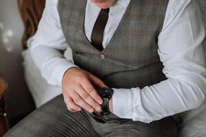 un de cerca de un recortado marco de un hombre pone en un reloj con un cuero cinturón, es vestido en un elegante traje, un blanco camisa, usa un oro anillo. foto