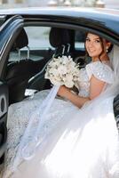 The bride is sitting in a black car with a bouquet of flowers in her hands and looks out of the car. Portrait of a rather shy bride in a car. Bride, smile, emotions. photo