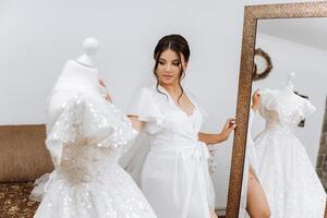 en el mañana, un increíblemente hermosa y lujoso novia, vestido en un blanco túnica, poses por el espejo en su dormitorio. Mañana de el novia con un lujoso peinado y natural constituir. foto