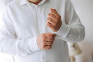 A man in a white shirt stands by the window in the room and fastens the buttons on his collar and sleeves. Watch on hand. Stylish business portrait of a man, close-up photo. The groom is preparing. photo