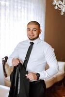 An elegant groom in a white shirt and a stylish tie is holding a jacket on a hanger. Details of the wedding morning in style. photo