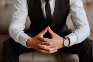 A close-up of a cropped frame of a man puts on a watch with a leather belt, is dressed in a stylish suit, a white shirt, wears a gold ring. photo