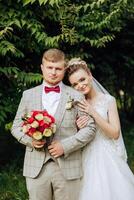 encantador y elegante recién casados son abrazando y sonriente en contra el antecedentes de otoño naturaleza en un hermosa jardín. un increíblemente hermosa joven novia inclinado en contra el hombro de su amado novio foto