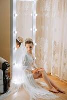 In the morning, an incredibly beautiful and luxurious bride, dressed in a white robe, poses by the mirror in her bedroom. MORNING of the bride with a luxurious hairstyle and natural makeup. photo