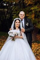 Stylish, young groom and beautiful bride in a long white dress and a long veil with a bouquet in their hands, hugging in the park in the autumn nature. Wedding portrait of newlyweds. photo