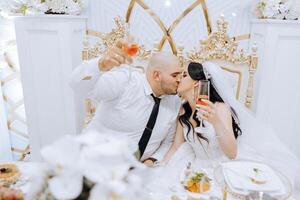 Young groom in a white shirt and smiling brunette bride at their table in a banquet hall, raising a toast and kissing against a background of fresh flowers. photo