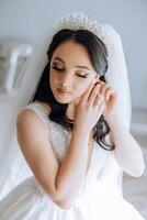 hermosa joven novia vistiendo pendientes antes de Boda ceremonia a hogar. un novia en un blanco Boda vestir pone en pendientes en su habitación en el Mañana. foto
