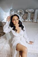 an incredibly beautiful bride in the morning in a white robe and a long veil sits on the bed in her bedroom. The bride poses in the morning before the wedding ceremony. photo