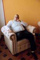 People, lifestyle, business, style, fashion and menswear concept. Positive successful young man sitting in a chair in a room, smiling at the camera, wearing elegant shoes, trousers, and a white shirt photo