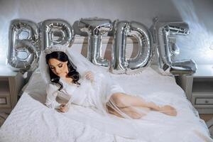 an incredibly beautiful bride in the morning in a white robe and a long veil lies on the bed in her bedroom near gel balls with the inscription bride. The bride poses in the morning. photo