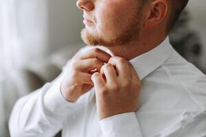 un hombre en un blanco camisa soportes por el ventana en el habitación y sujeta el botones en su collar y mangas reloj en mano. elegante negocio retrato de un hombre, de cerca foto. el novio es preparando. foto