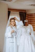 portrait of an incredibly beautiful girl bride in a white robe with a glass of champagne in her hand next to her wedding dress. The bride poses in the morning before the wedding ceremony. photo