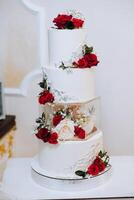 A large luxurious multi-tiered wedding cake is decorated with fresh red rose flowers in the banquet hall. Wedding dessert under the evening light. Wedding decor. photo