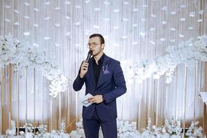 Handsome young presenter in dark blue suit holding wireless microphone and clipboard while standing on photo booth while waiting for wedding ceremony.