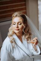 portrait of an incredibly beautiful girl bride in a white robe with a glass of champagne in her hand in the bedroom. The bride poses in the morning before the wedding ceremony. photo