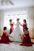 Bridesmaids rejoice in the morning, helping to fasten the buttons on the wedding dress and prepare for the wedding ceremony. They take pictures, smile, help the bride with her shoes. photo