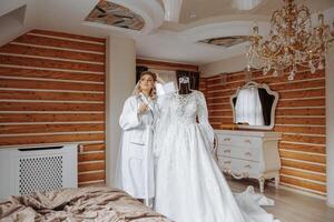 portrait of an incredibly beautiful girl bride in a white robe with a glass of champagne in her hand next to her wedding dress. The bride poses in the morning before the wedding ceremony. photo