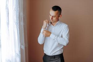 A man in a white shirt stands by the window in the room and fastens the buttons on his collar and sleeves. Watch on hand. Stylish business portrait of a man, close-up photo. The groom is preparing. photo