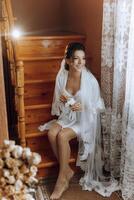 portrait of an incredibly beautiful bride girl in a white robe, sitting on wooden stairs in the hallway, the bride poses in the morning before the wedding ceremony. photo