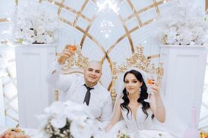 A young groom in a white shirt and a smiling brunette bride are at their table in a banquet hall, raising a toast against a background of fresh flowers. photo