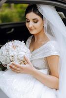 Wedding portrait. A brunette bride in an elegant dress and veil poses with a bouquet in the car. Preparation for the celebration. Beautiful hair and makeup. photo