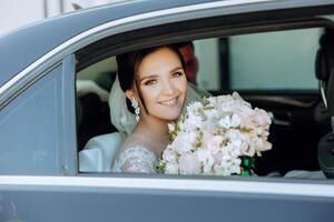 el novia es sentado en un negro coche con un ramo de flores de flores en su manos y mira fuera de el coche. retrato de un bastante tímido novia en un coche. novia, sonrisa, emociones foto