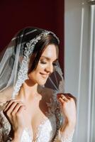 Beautiful young bride holding veil in white wedding dress, portrait of brunette bride in hotel room, morning before wedding. photo