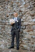 retrato de un hermoso joven novio en un otoño día al aire libre en un elegante traje y participación un Boda ramo de flores de flores al aire libre foto. hermoso hombre en un negocio traje. foto