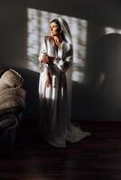 portrait of an incredibly beautiful girl bride in a white robe in the bedroom in beautiful light and shadow, the bride poses in the morning before the wedding ceremony. photo