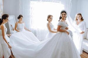 Bridesmaids rejoice in the morning, helping to fasten the buttons on the wedding dress and prepare for the wedding ceremony. They take pictures, smile, help the bride with her shoes. photo