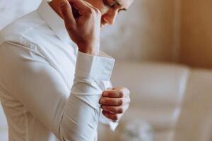 un hombre en un blanco camisa soportes por el ventana en el habitación y sujeta el botones en su collar y mangas reloj en mano. elegante negocio retrato de un hombre, de cerca foto. el novio es preparando. foto