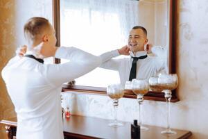 un joven hombre en un blanco camisa soportes por el ventana en el habitación y pone en un atar. el novio obtiene vestido en el Mañana y prepara para el boda. foto