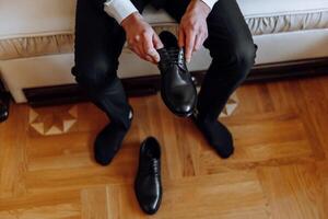 A man is putting on his shoes while sitting on a sofa in a hotel room. A young entrepreneur is preparing to go on a business trip. Businessman preparing for travel in home bedroom early in the morning photo