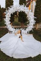 un morena novia en un velo y un tiara en su pelo se sienta en un untado vestido, posando cerca un blanco arco hecho de flores en el forma de un círculo. en un blanco antecedentes. soleado día. Boda ceremonia foto